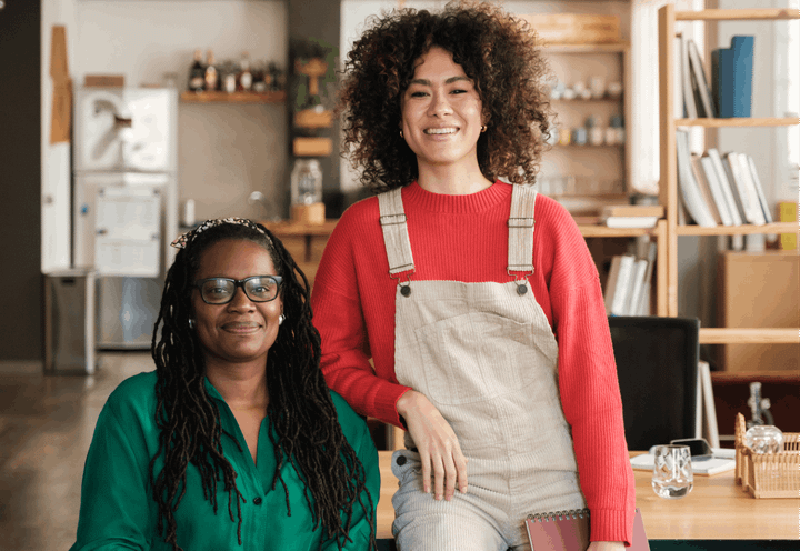 Two women smiling together one is seated in a green shirt and the other is leaning on her in a red jumper and cream dungarees.