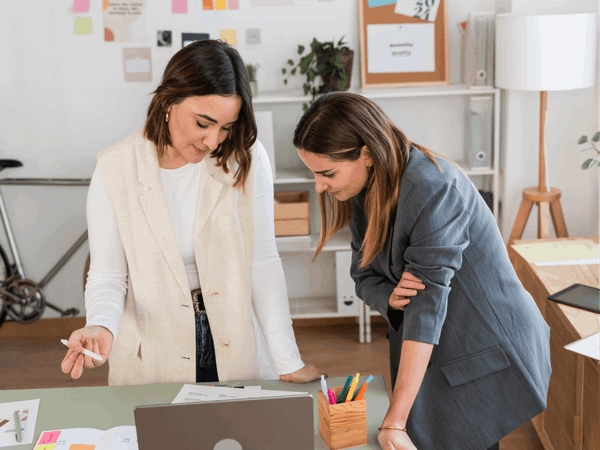 Two women are standing in a modern office together and reading off a page.