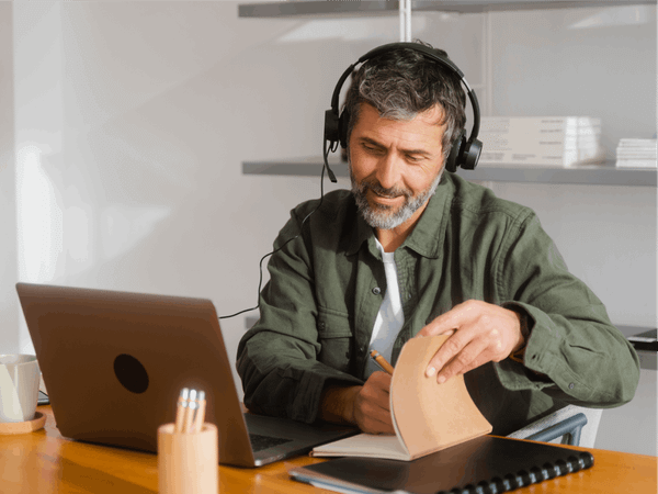 A man is sat at his laptop with headphones on and taking notes.
