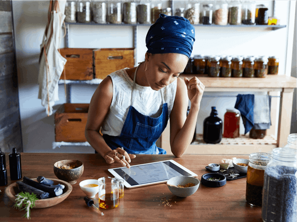 A woman is in a kitchen, wearing her apron whilst reading off a tablet.