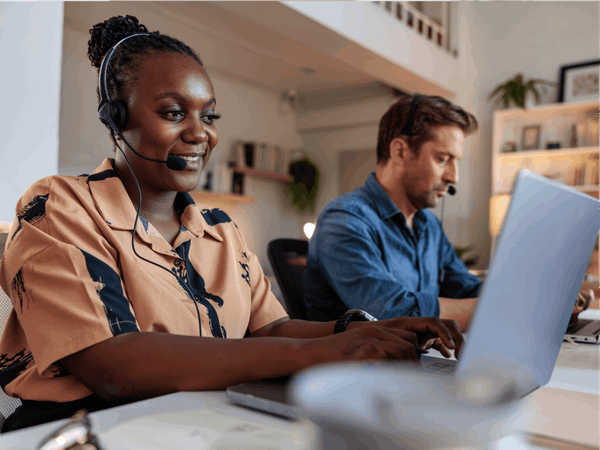 A woman is sat next to a man they are both using laptops with headphones on.