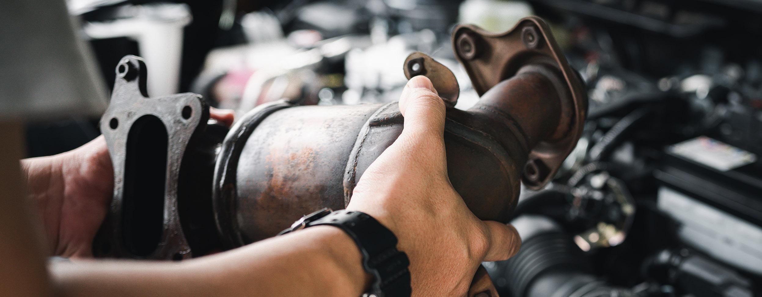 Nahaufnahme eines alten Katalysators in der Hand eines Mechanikers. Im Hintergrund ist der Motorraum zu sehen.