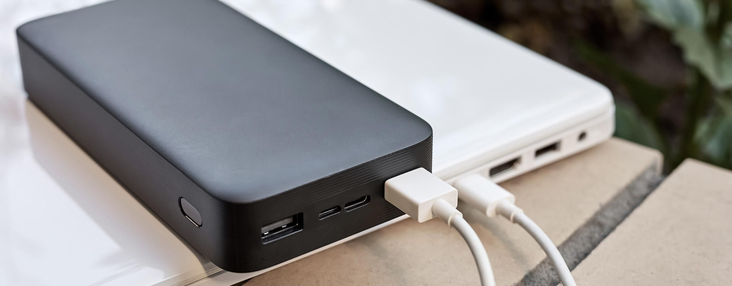 Ein weißer Laptop liegt auf einer Mauer und ist über ein USB-Kabel mit einer schwarzen Powerbank verbunden.