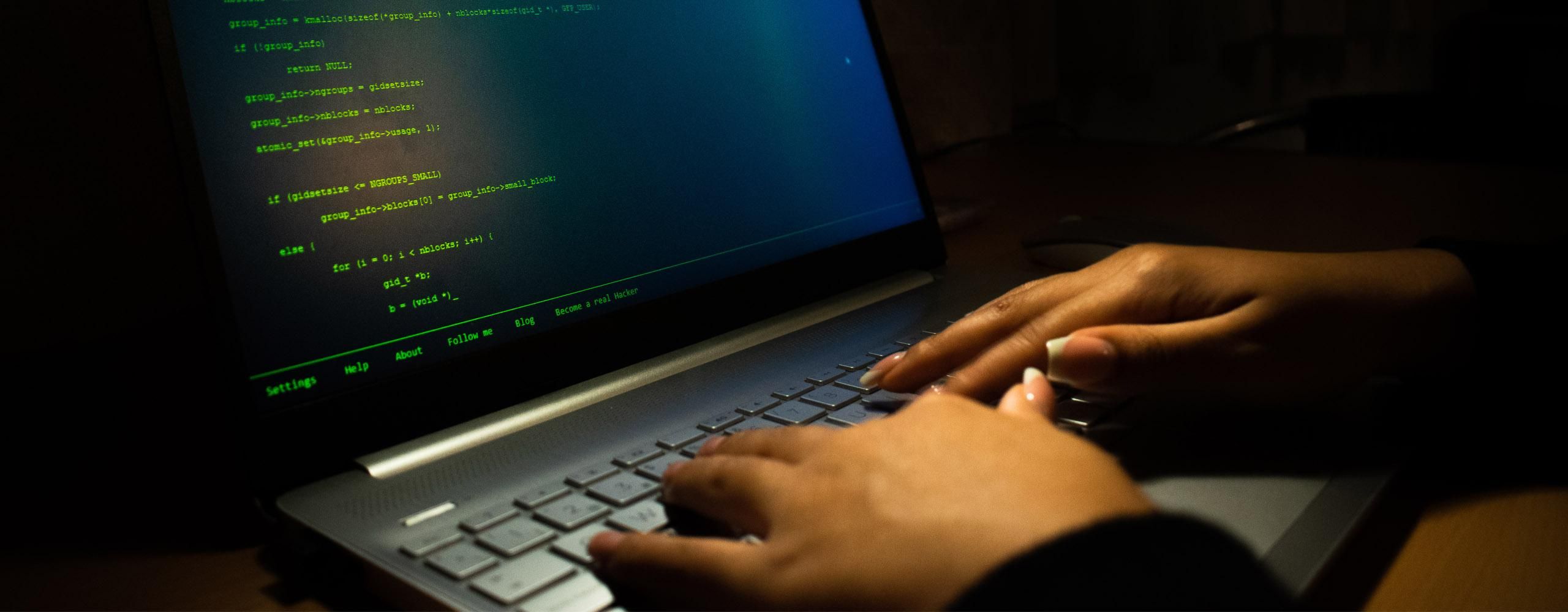 Nahaufnahme eines Laptops, auf dem eine Person mit ihren Händen tippt. Auf dem ansonsten dunklen Bildschirm ist Code einer Programmiersprache zu sehen.