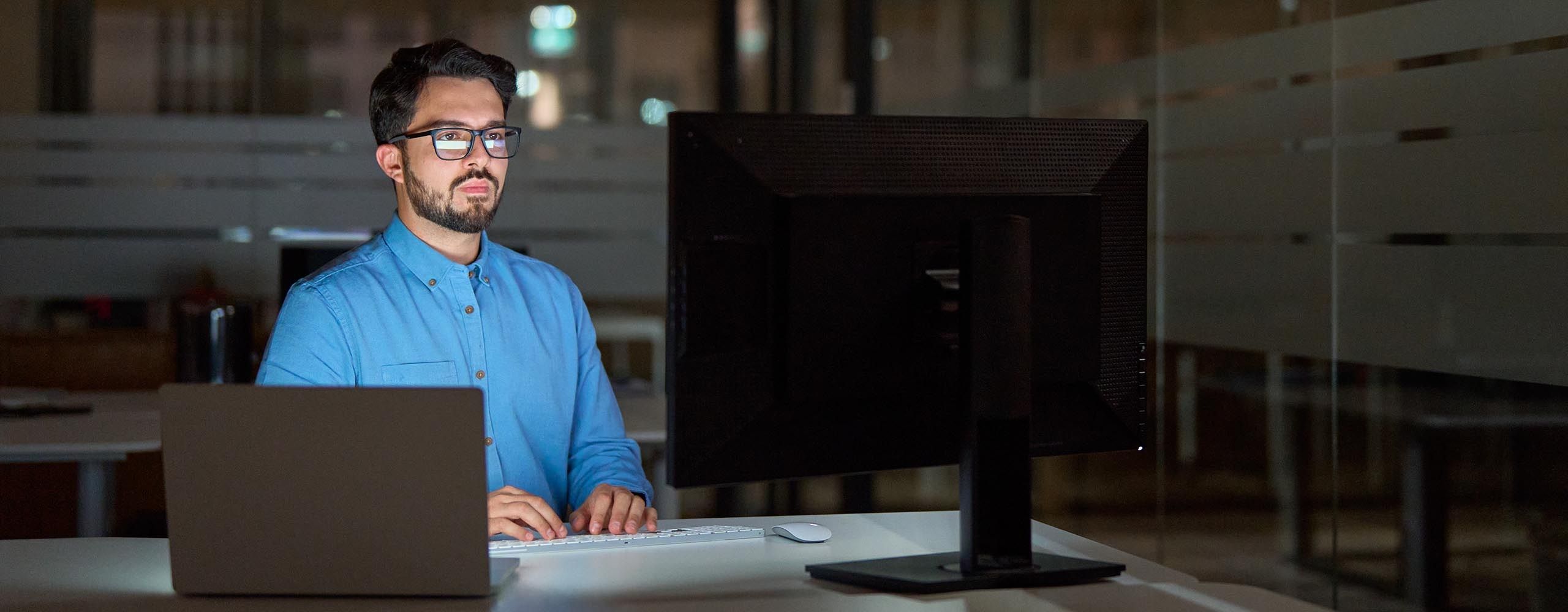 Eine Person mit blauem Hemd und in gerader Haltung sitzt an einem Laptop und schaut auf einen Monitor.